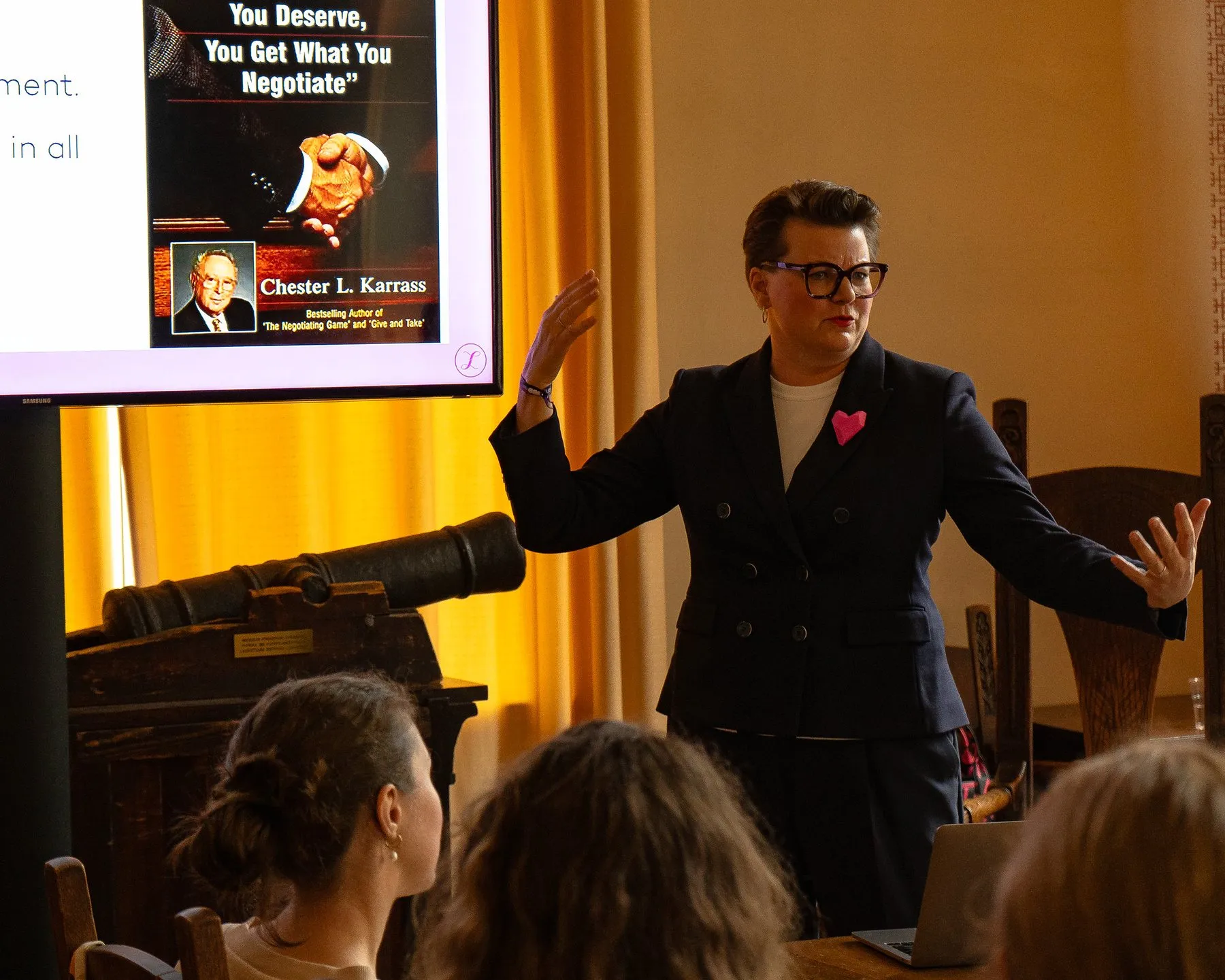 A speaker in a dark suit addressing a seated audience beside a projected slide
