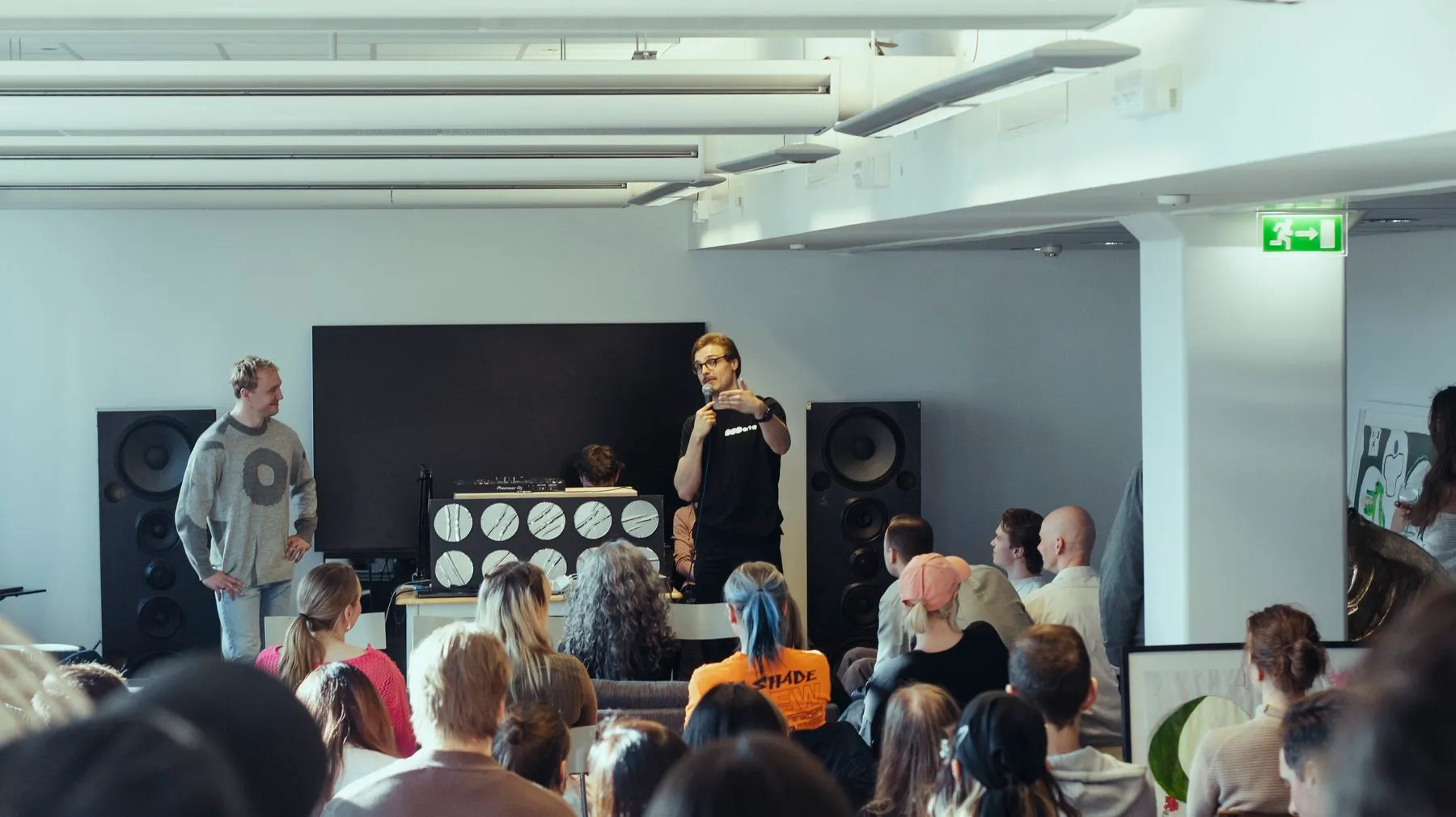 A talk in front of the DJ booth and speakers, seen from the back of the audience