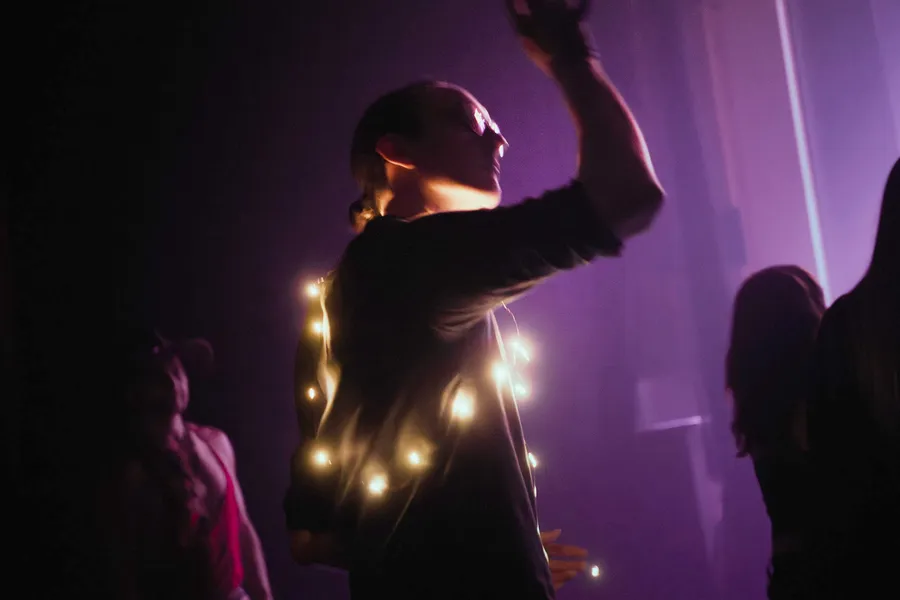 A performer in silhouette with one arm raised, strings of lights behind