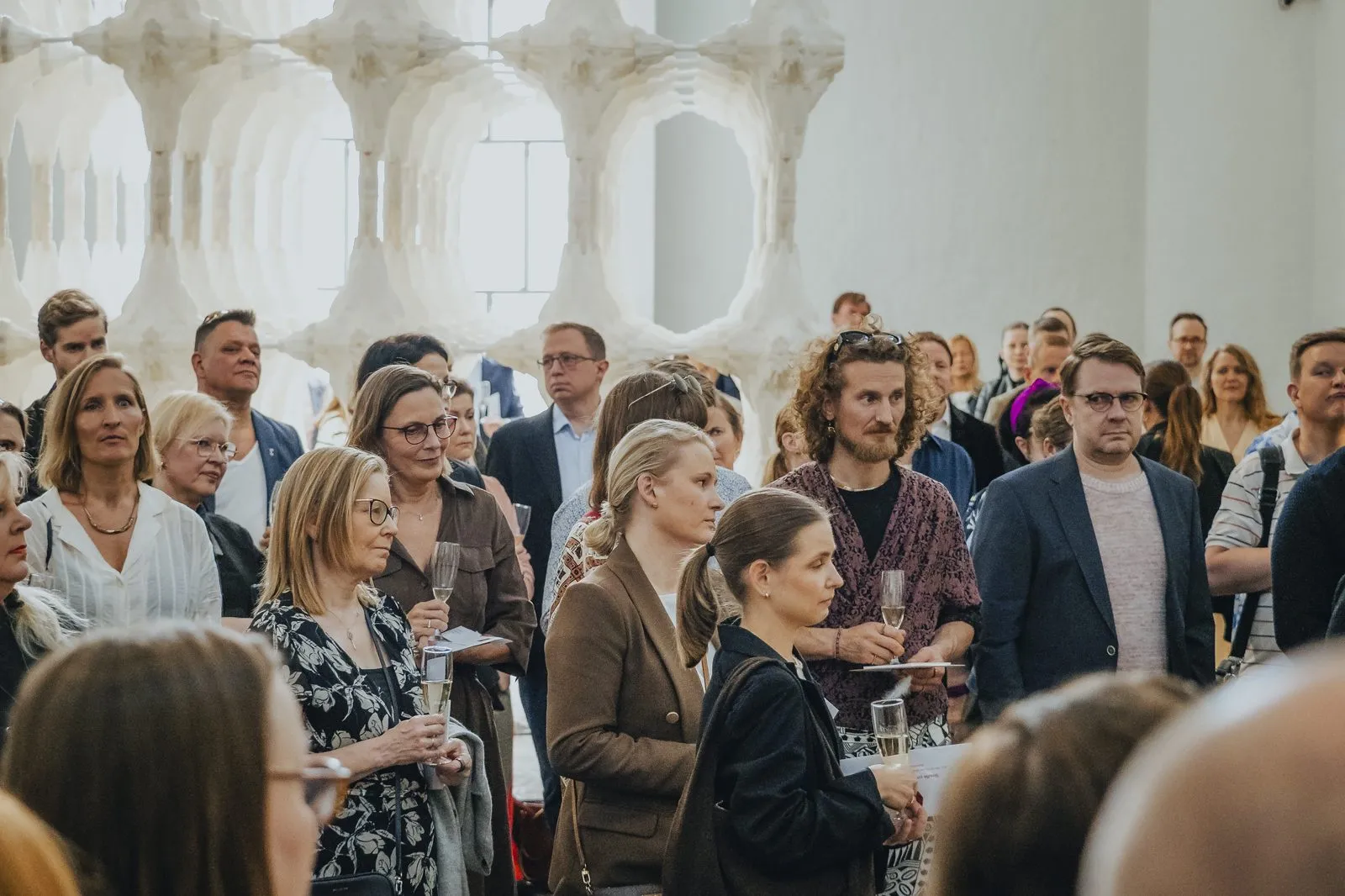 A standing crowd listening in a white-walled hall